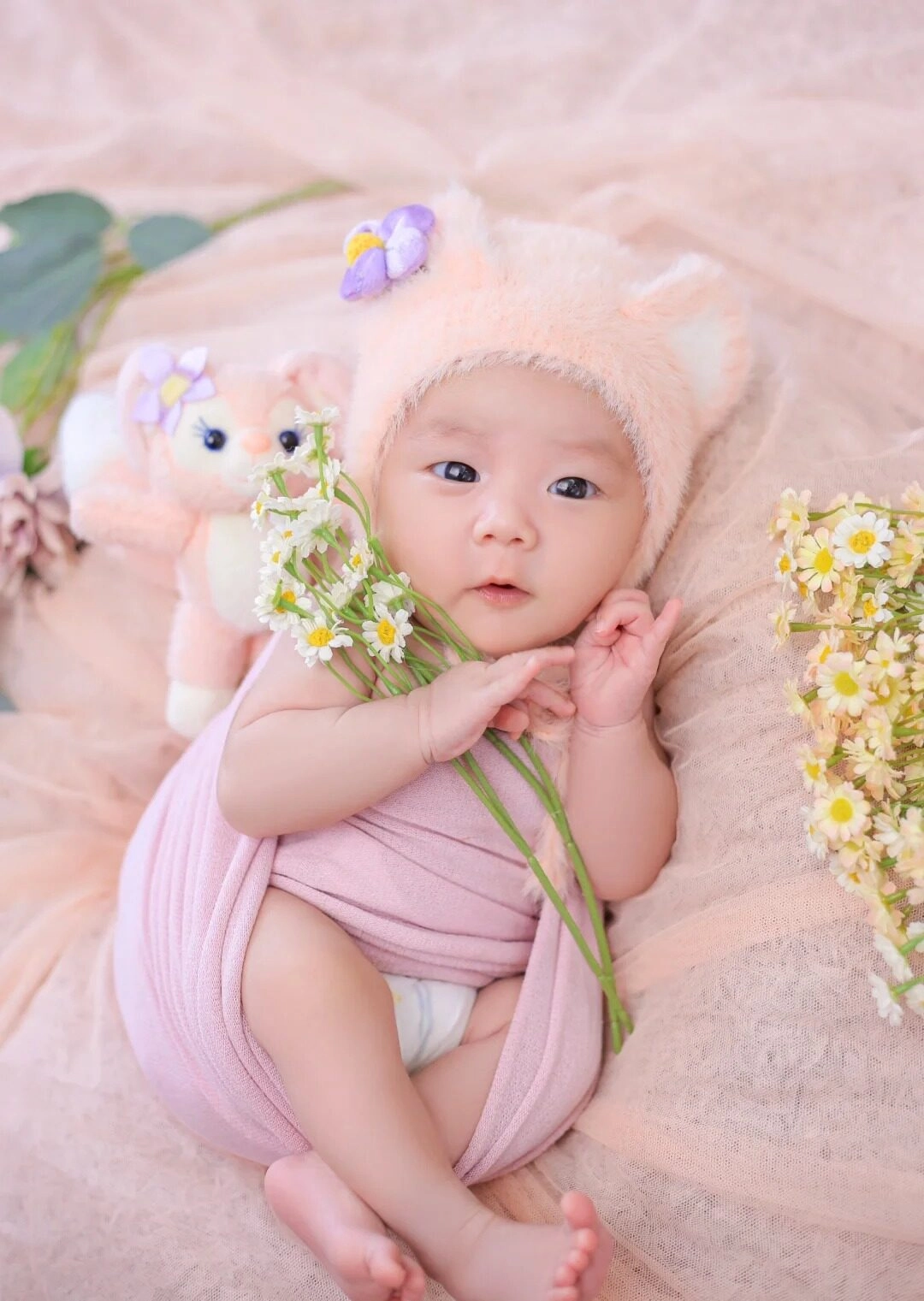 Bé gái quấn khăn hồng, cầm cành hoa nhỏ, ánh mắt ngây thơ đáng yêu.