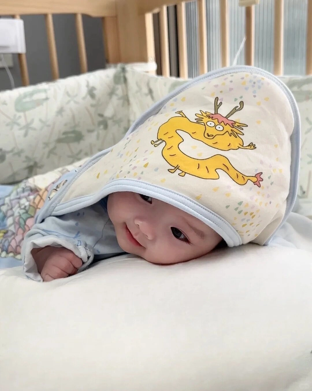 Bé nằm sấp trong chăn, nụ cười lém lỉnh như đang chơi trốn tìm.