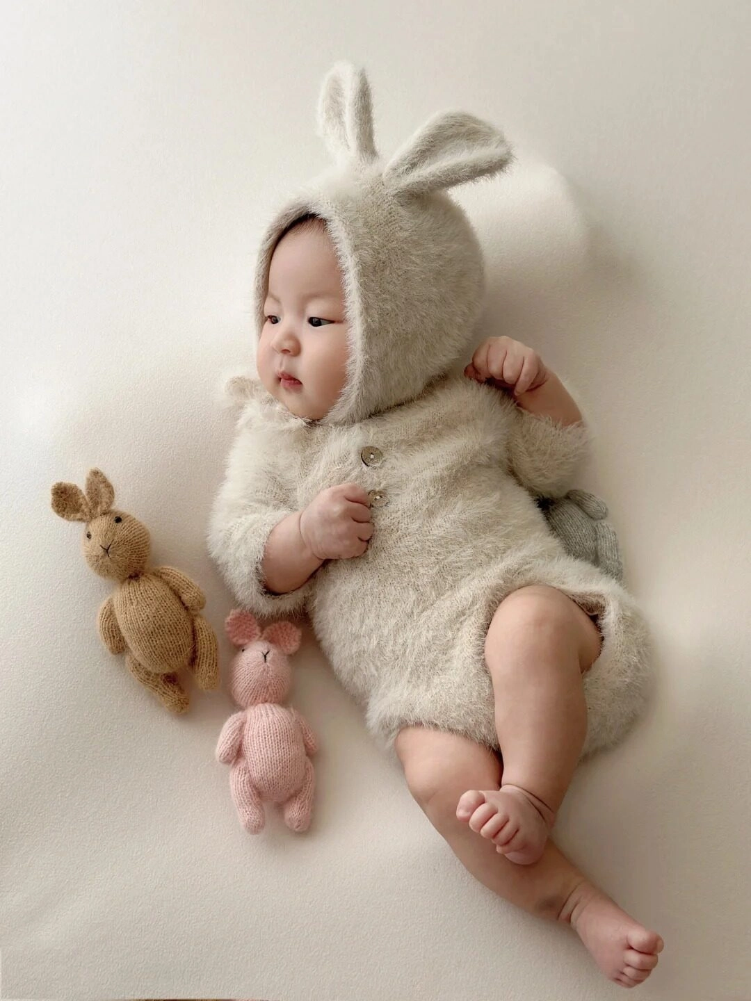 Bé diện bộ đồ thỏ bông, nằm cạnh hai bạn thỏ nhỏ đáng yêu.