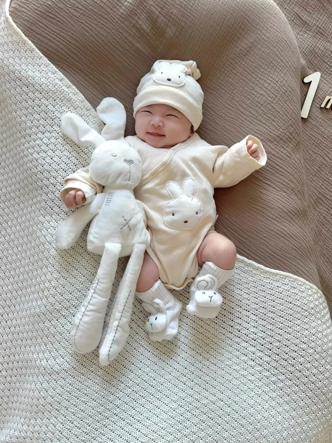 Bé mặc đồ trắng với họa tiết thỏ, cười rạng rỡ kỷ niệm một tháng tuổi.