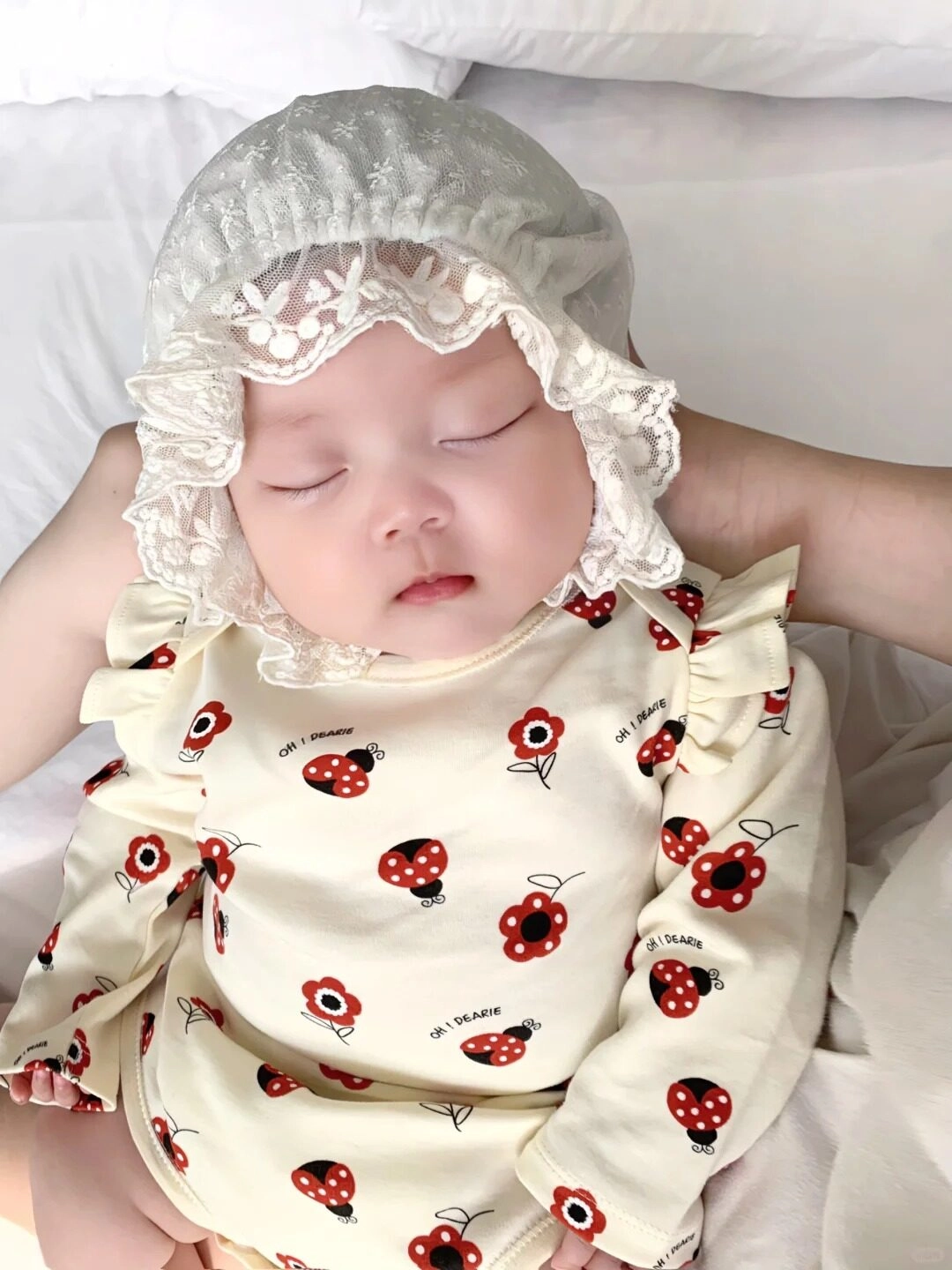 Bé ngủ ngoan trong váy chấm bi đỏ, mũ ren trắng tạo nét cổ điển.