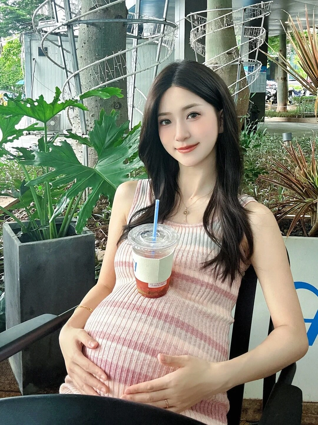 Váy hồng ôm dáng, mẹ bầu tựa vào ghế ngoài trời, ly trà đặt hờ trên bụng.