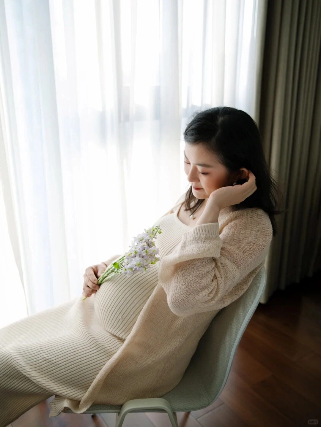 Váy len kem dài, dáng ngồi thư thái bên cửa sổ, tay ôm hoa nhỏ dịu dàng.