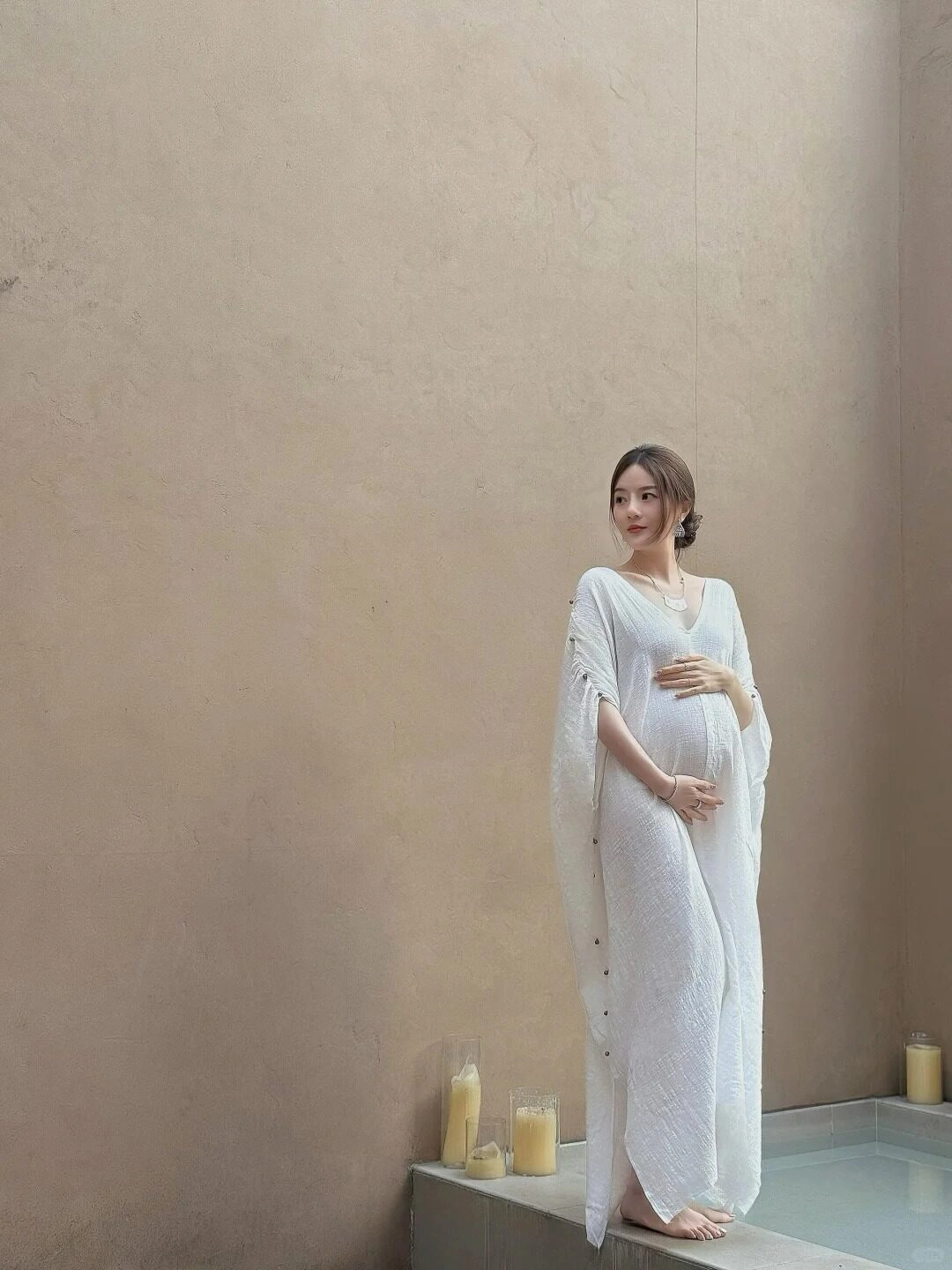 Cô gái diện váy trắng suông nhẹ, tay đặt lên bụng bầu, ánh mắt trầm tư.