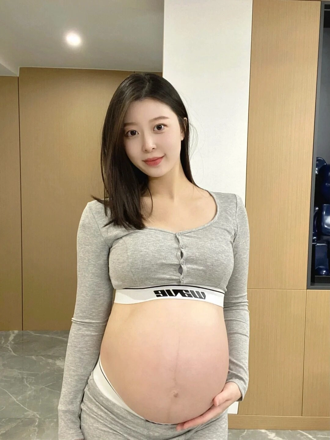 Váy body xám dài tay, phần bụng bầu nổi bật, nụ cười rạng rỡ.