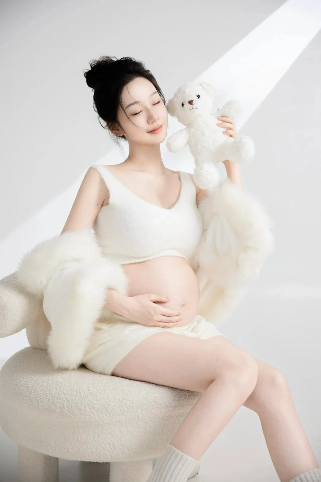 Bộ đồ lông trắng mềm mại, mẹ bầu nâng niu gấu bông, ánh mắt tràn đầy yêu thương.