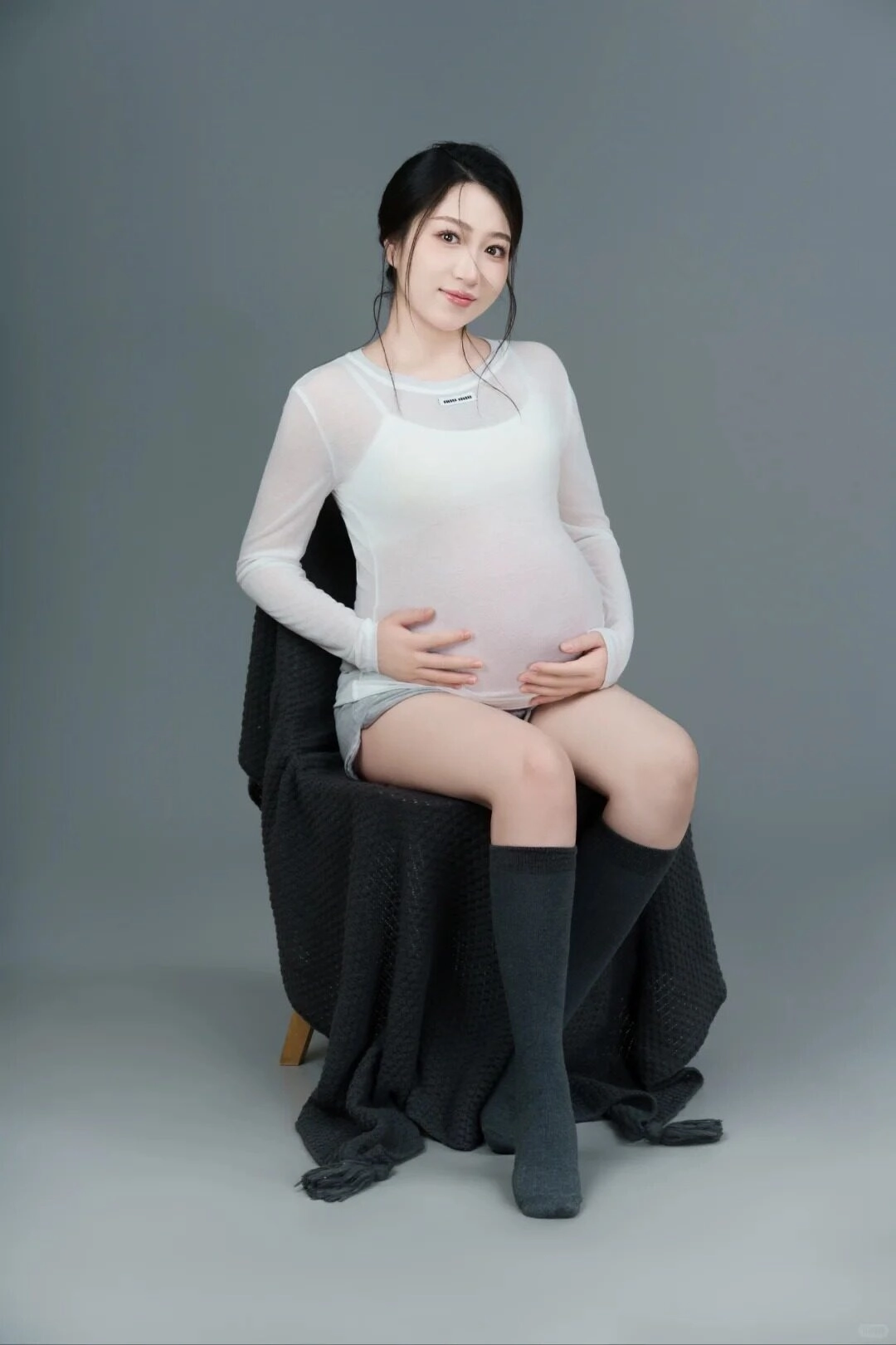 Ngồi thư giãn trên ghế, mẹ bầu mặc áo trắng và quần short xám, đầy dịu dàng.