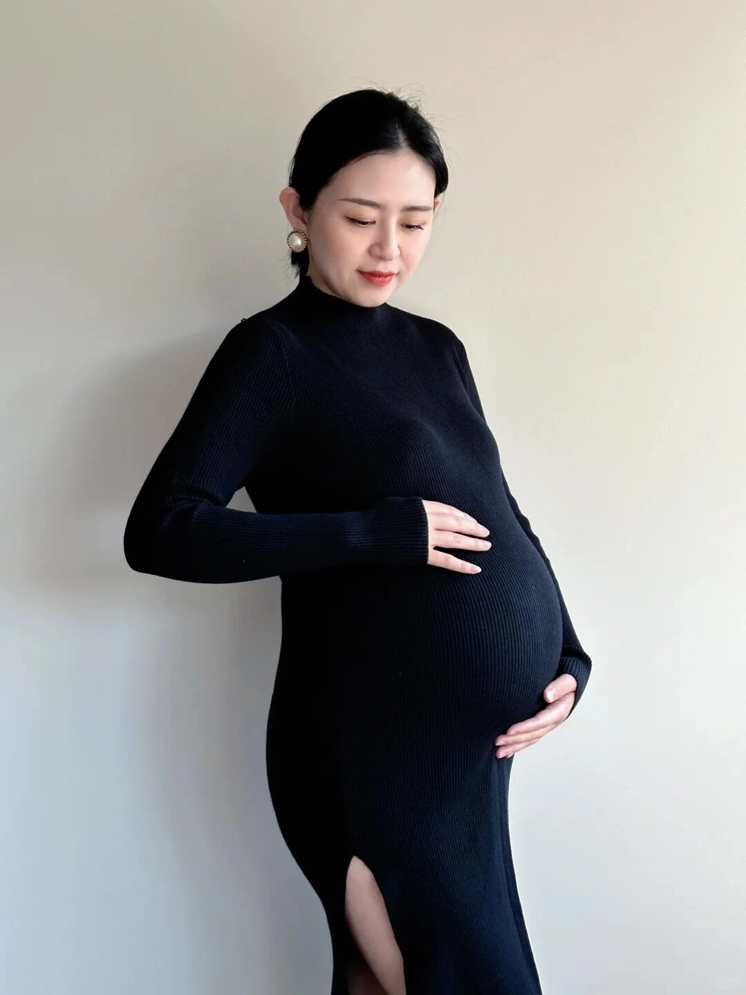 Váy len đen ôm sát, mẹ dịu dàng vuốt ve bụng bầu trong ánh sáng nhẹ.