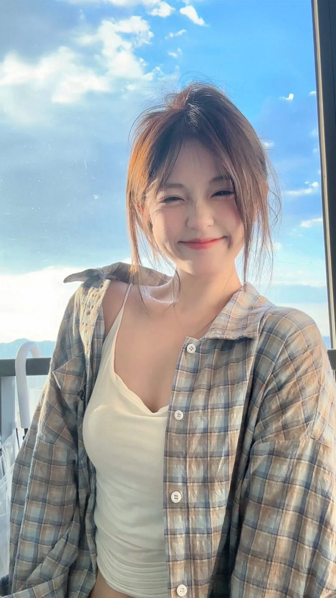 Kiểu tóc buộc thấp nhẹ nhàng, áo hai dây trắng phối sơ mi caro khoác hờ.