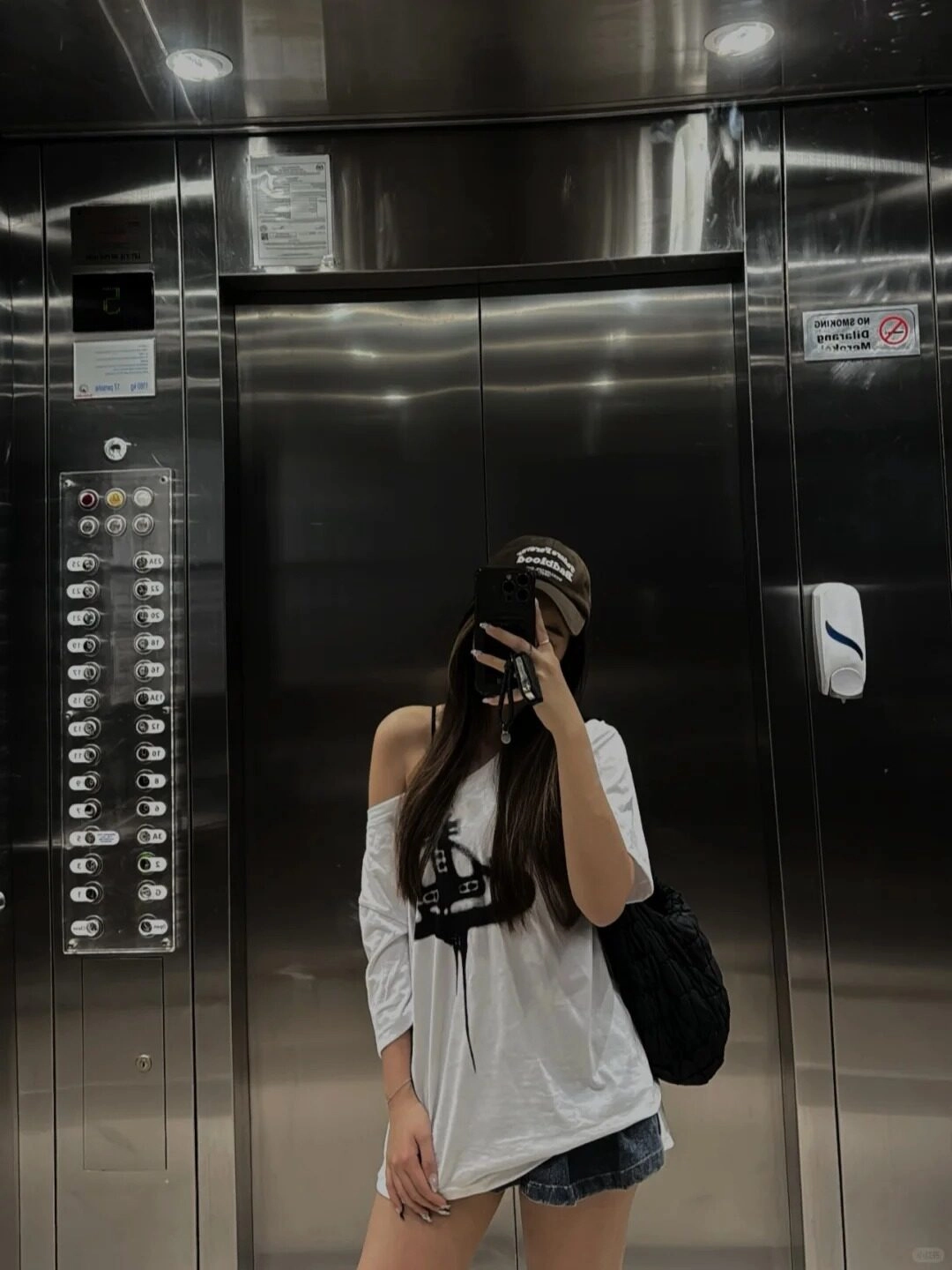 Áo trắng vai, quần short jean, đội mũ, tạo kiểu dáng trong thang máy.