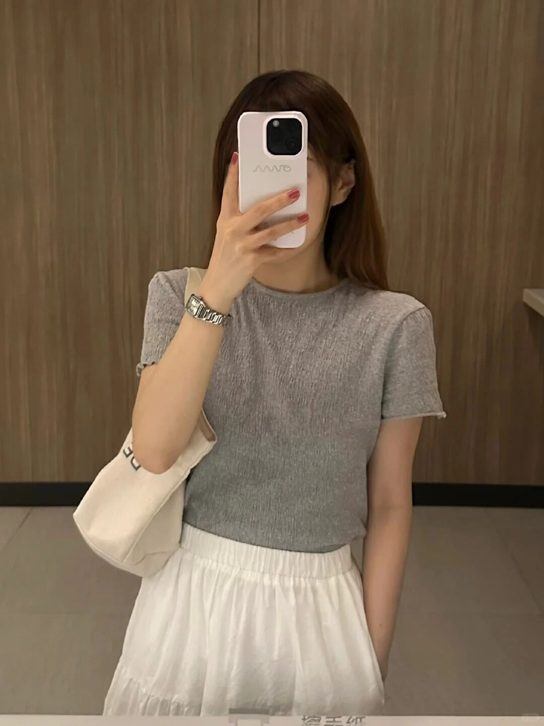 Áo thun xám, chân váy trắng, tay cầm điện thoại che kín khuôn mặt selfie.