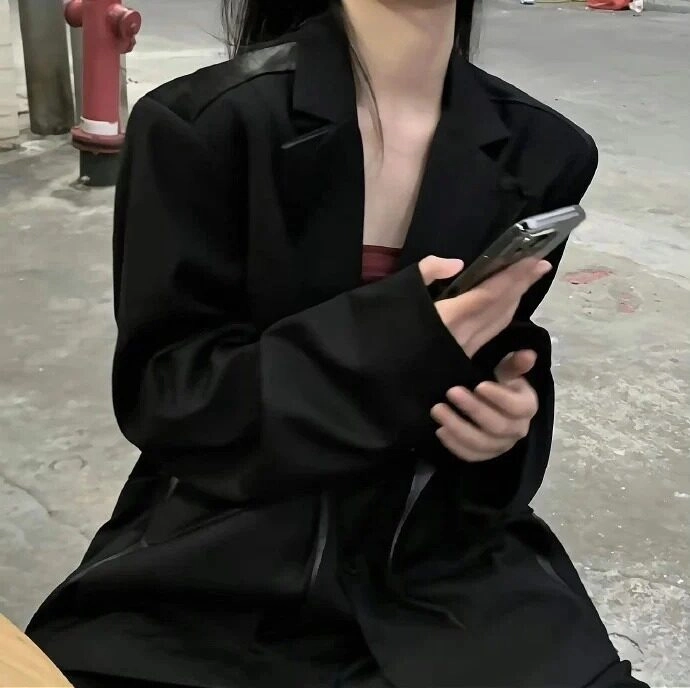 Ngồi trên vỉa hè, diện áo khoác đen, tay cầm điện thoại, vibe lạnh lùng.