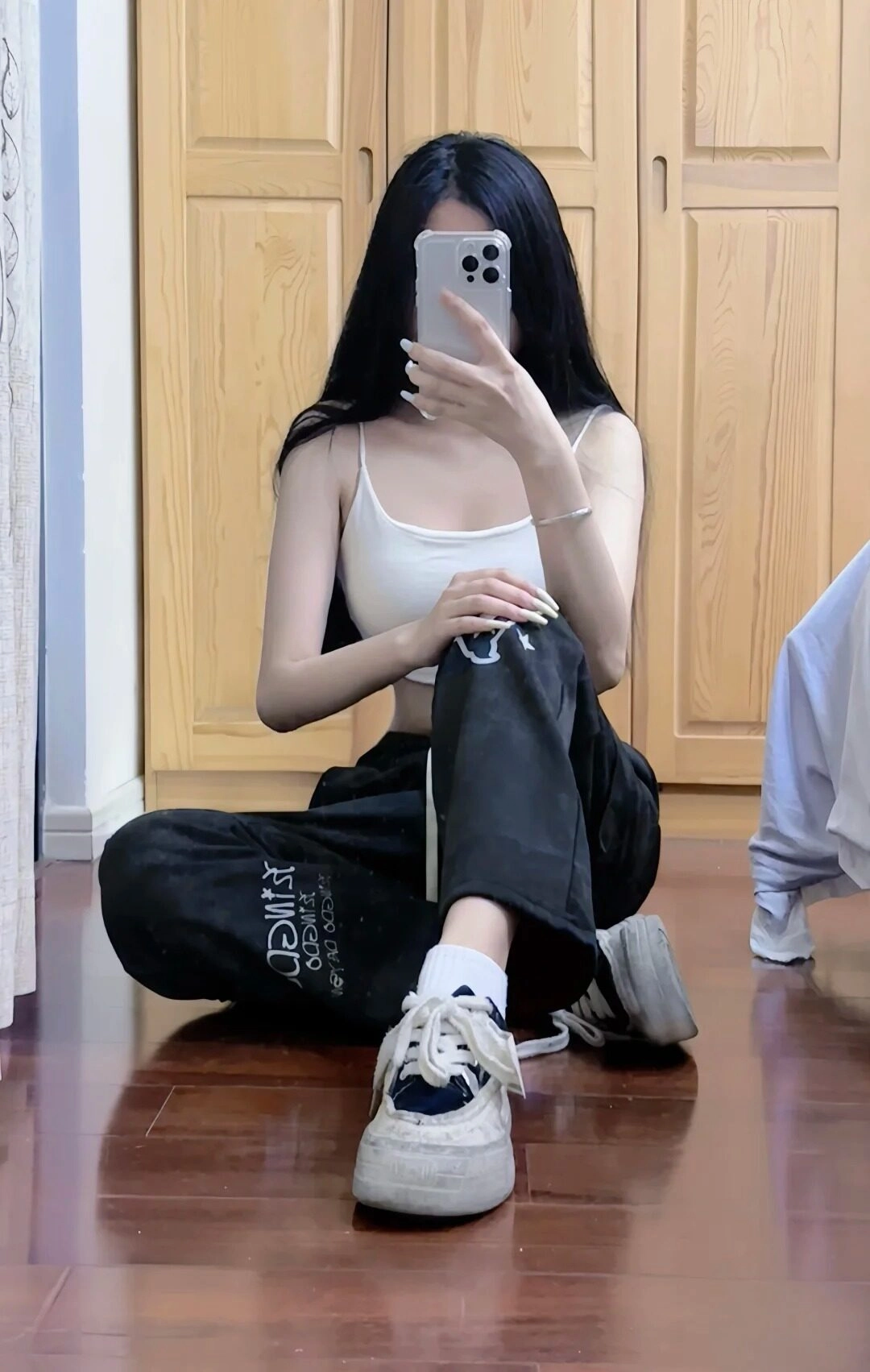 Ngồi trên sàn, che mặt bằng điện thoại, diện áo croptop trắng và quần baggy đen.