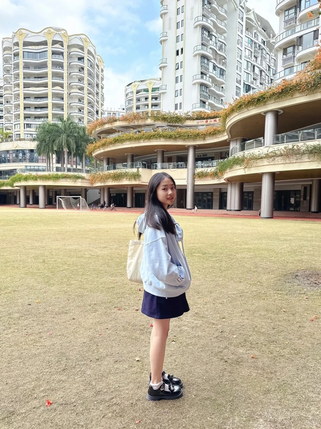 Nữ sinh đứng giữa sân trường rộng, khung cảnh hiện đại phía sau.