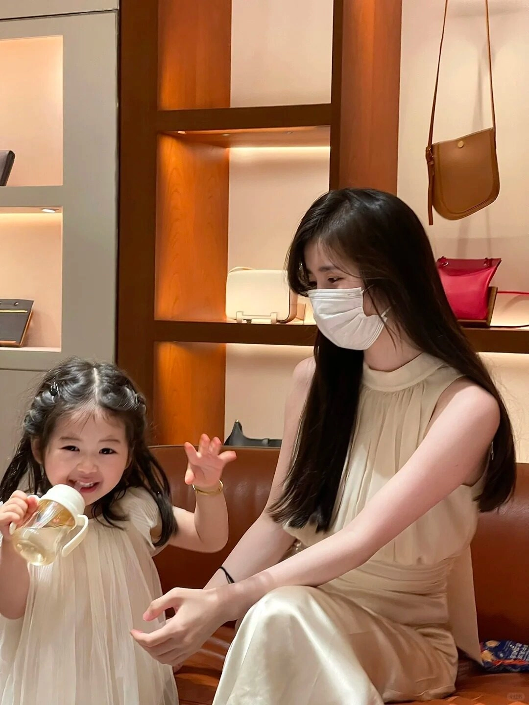 Mẹ mặc váy kem thanh lịch, bé cầm bình sữa cười tươi, khoảnh khắc ấm áp.