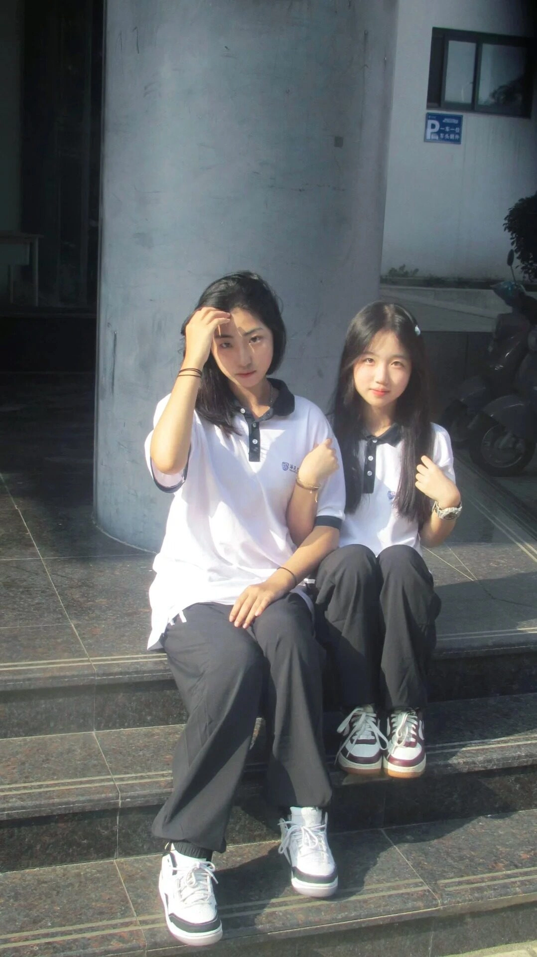 Đôi bạn gái ngồi tầng lớp, tóc ngang vai tự nhiên, trang phục sinh khỏe mạnh.