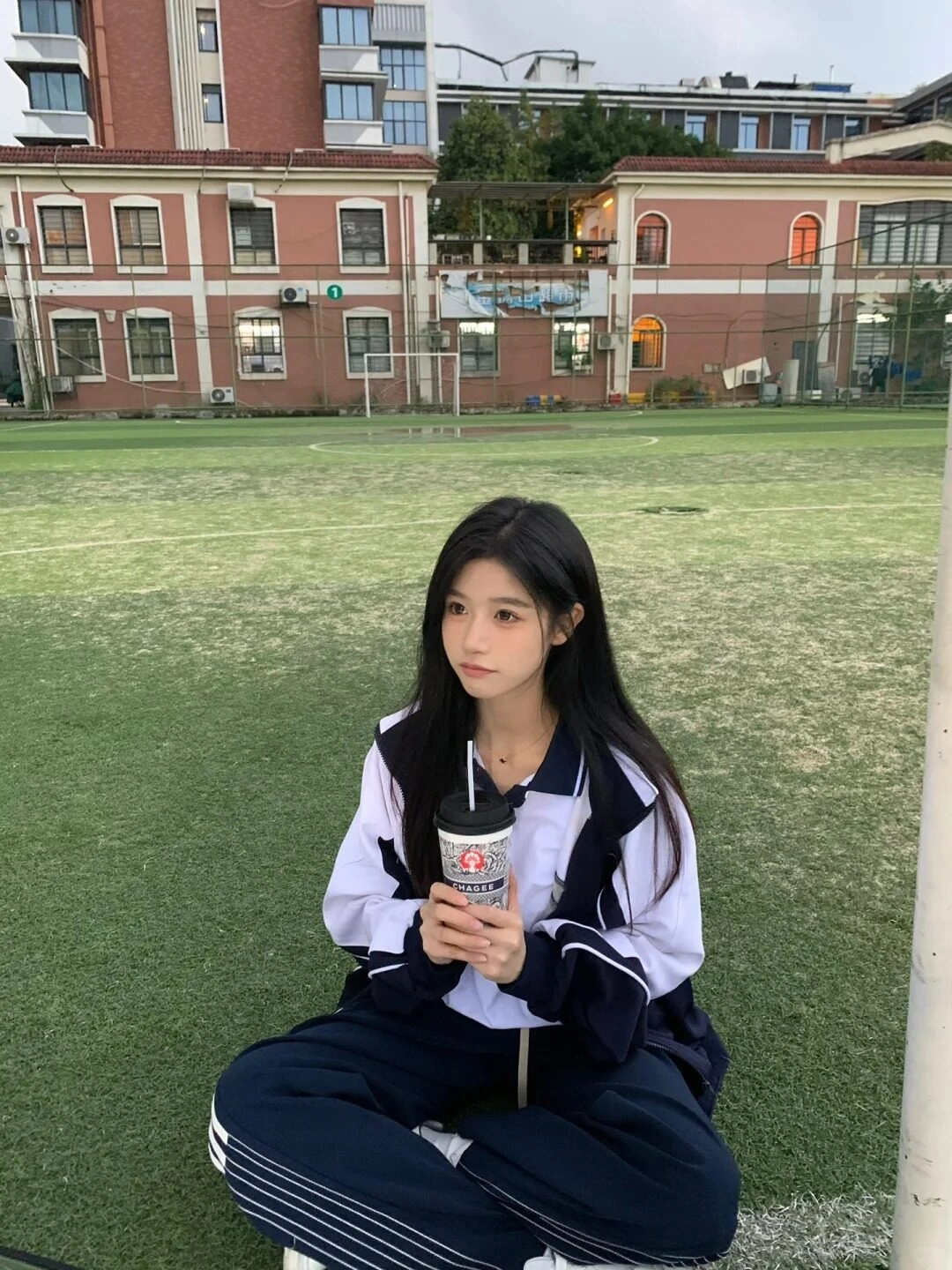 Cô bạn tóc dài ngồi sân bóng, trang phục thể thao, tay cầm cốc nước nước.