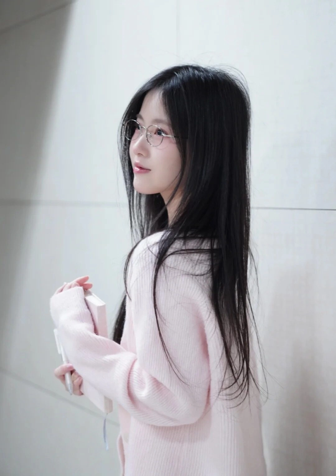 Thiếu nữ áo len hồng, mái tóc dài đen tạo dáng dễ thương bên tường học.