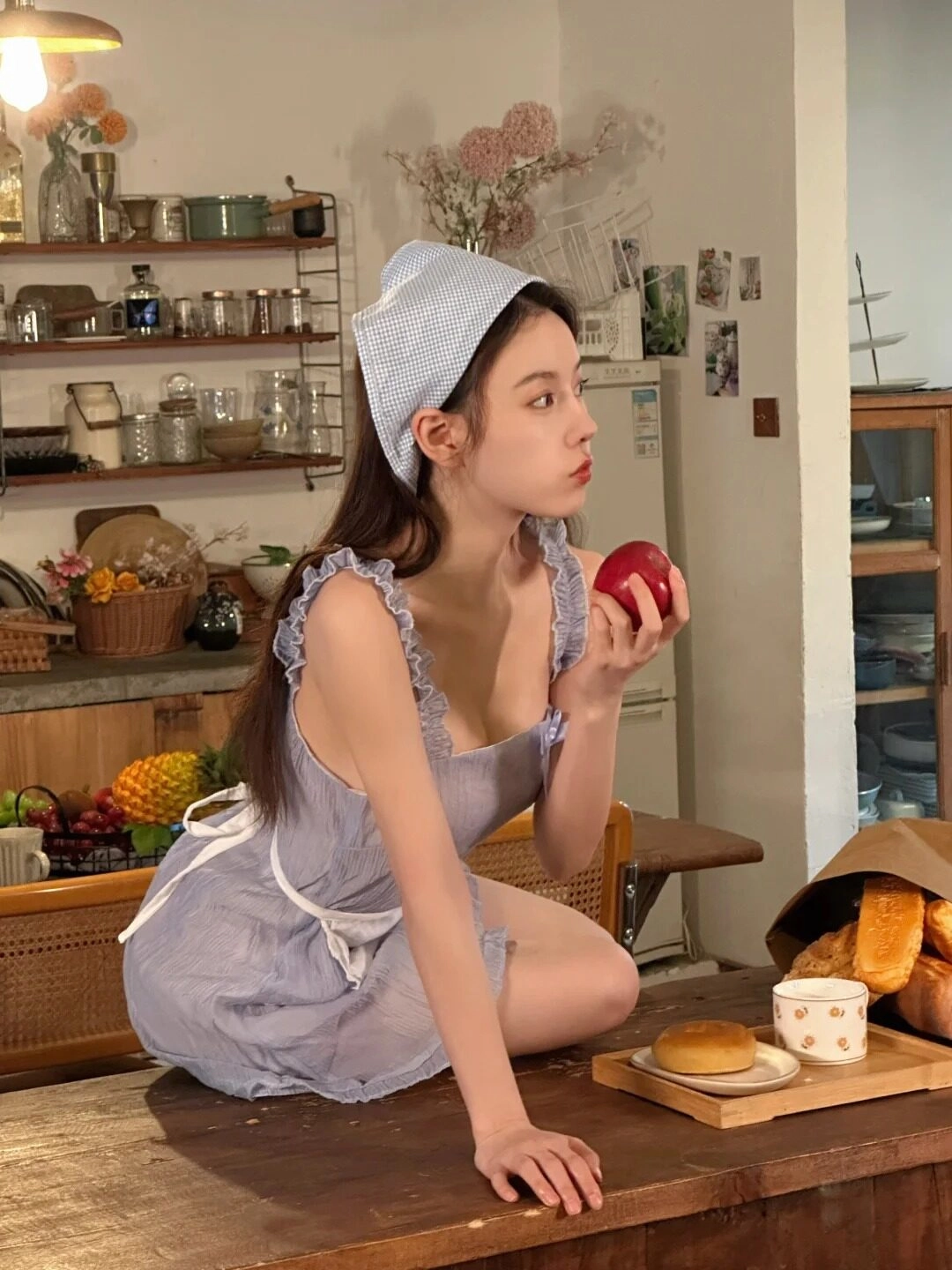Thiếu nữ mặc váy xanh tím nhẹ, tóc dài cài khăn, ngồi ăn táo trong bếp.
