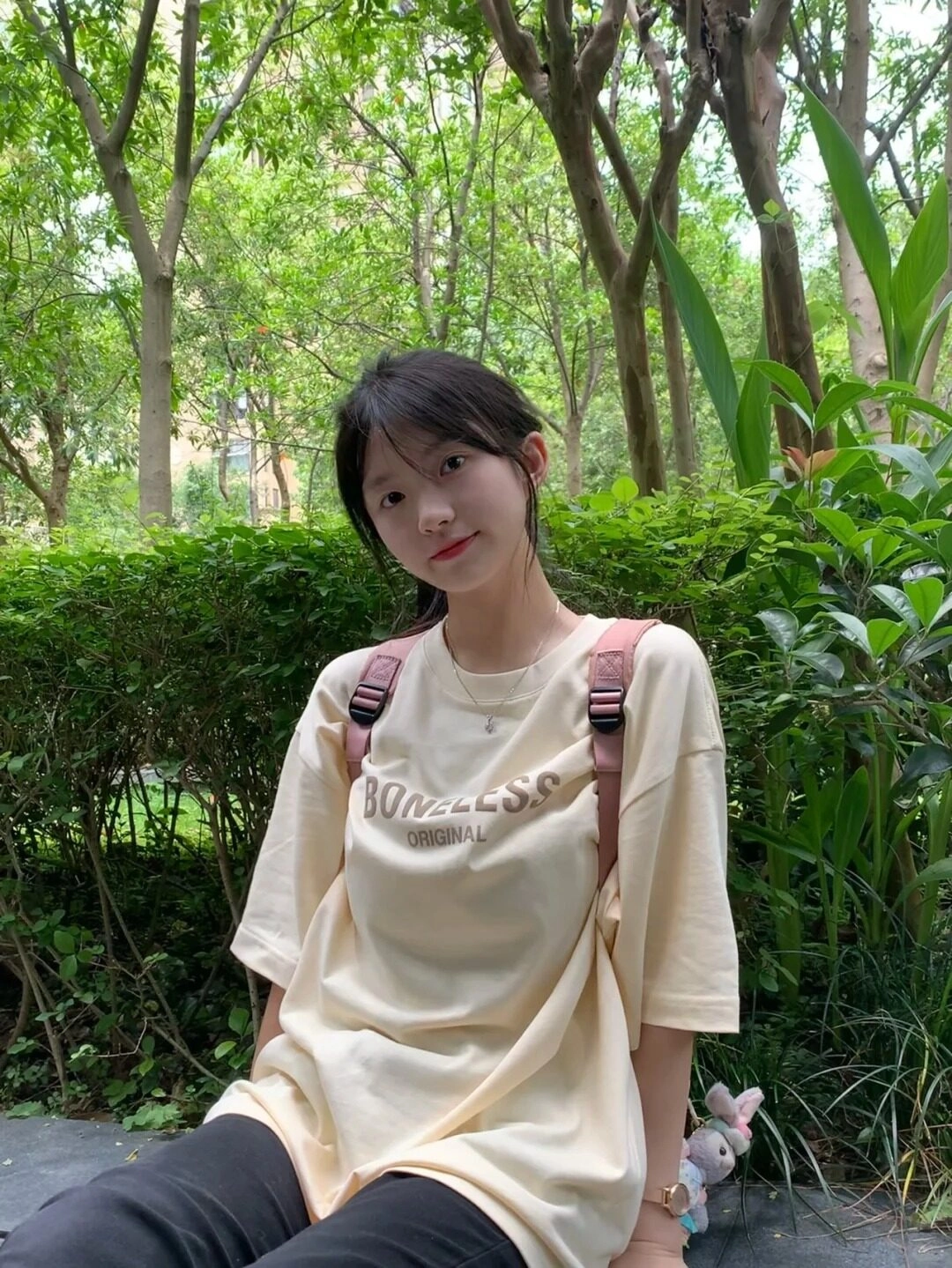 Nữ sinh 2k10 áo nữ thun vàng nhạt, tóc cột gọn, ngồi thư giãn giữa vườn cây.